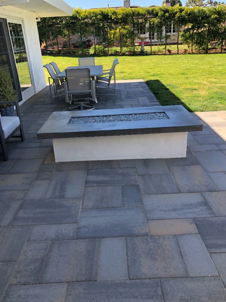 Linear white and charcoal gas fire pit with blue fire glass on a gray paver patio beside outdoor dining