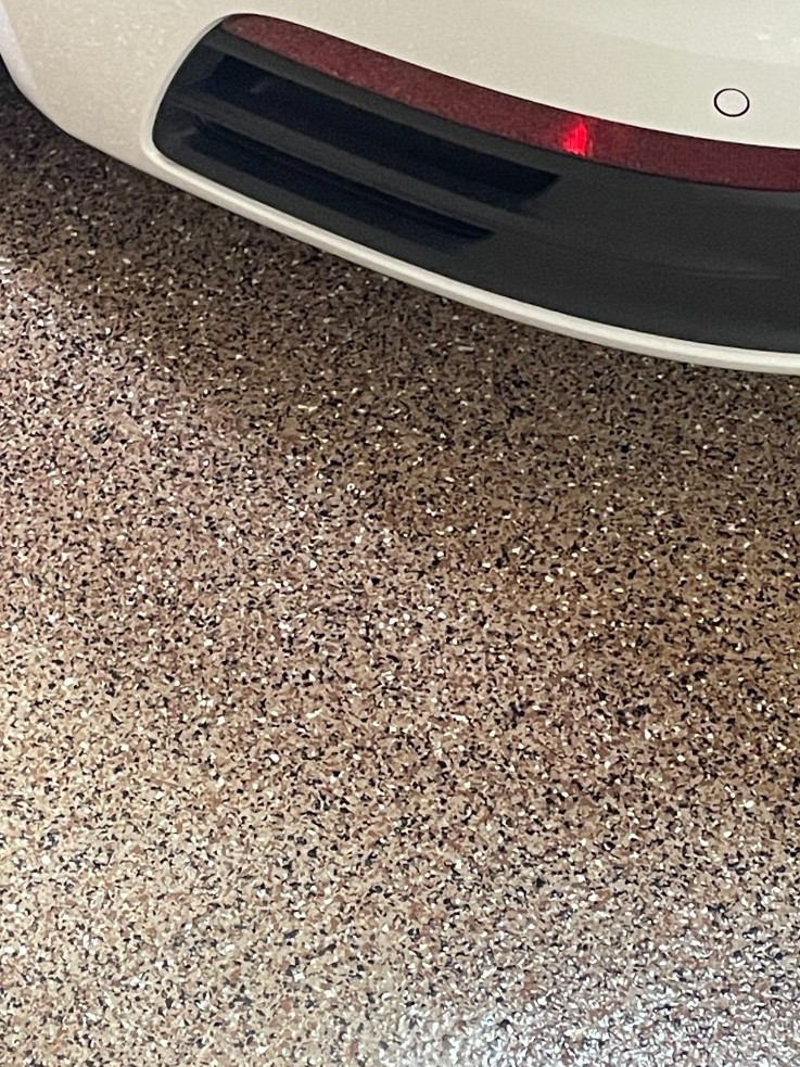 Close-up of glossy tan and brown flake epoxy garage floor beside a white car bumper