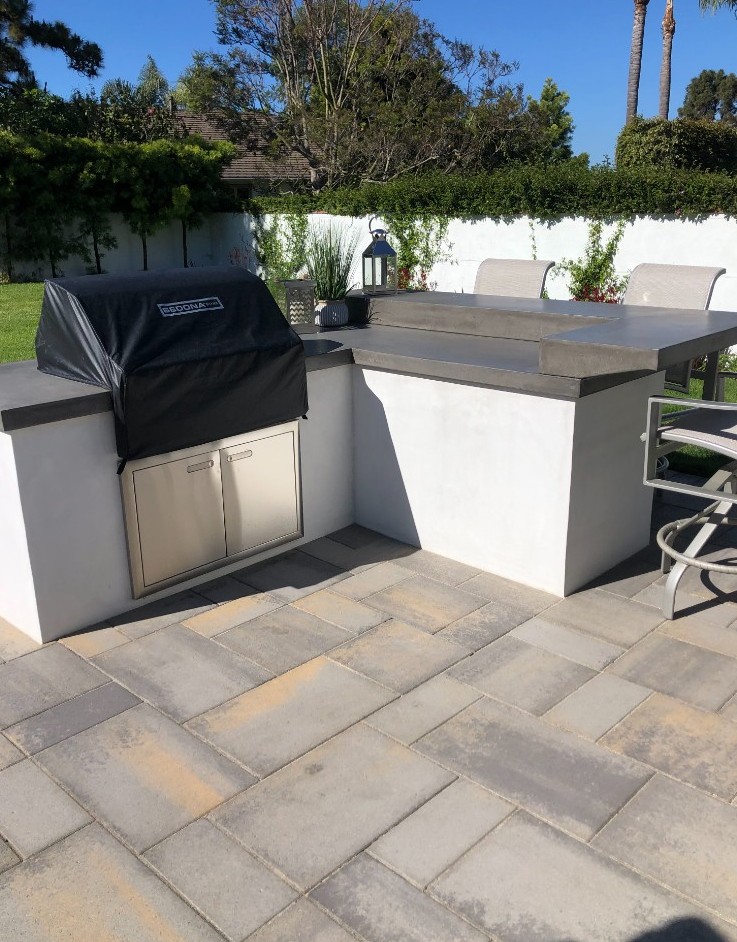White outdoor kitchen island with concrete counters and bar stools on a multicolor paver patio