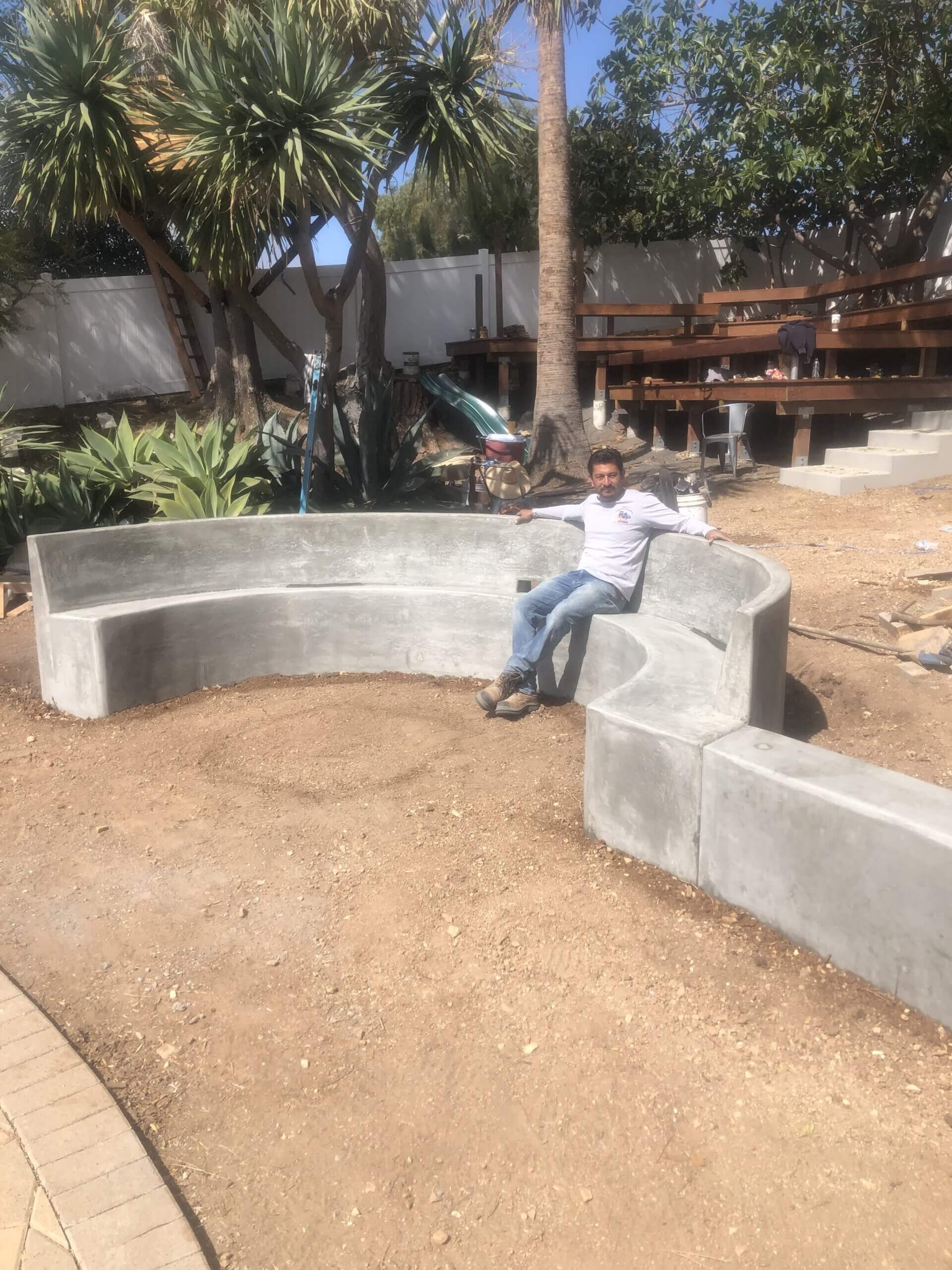 A crew member sitting on a freshly formed curved concrete bench in a backyard mid-construction