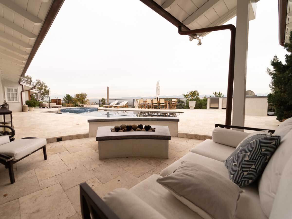 Covered lounge with a fire table on a travertine pool deck, spa and hilltop view beyond