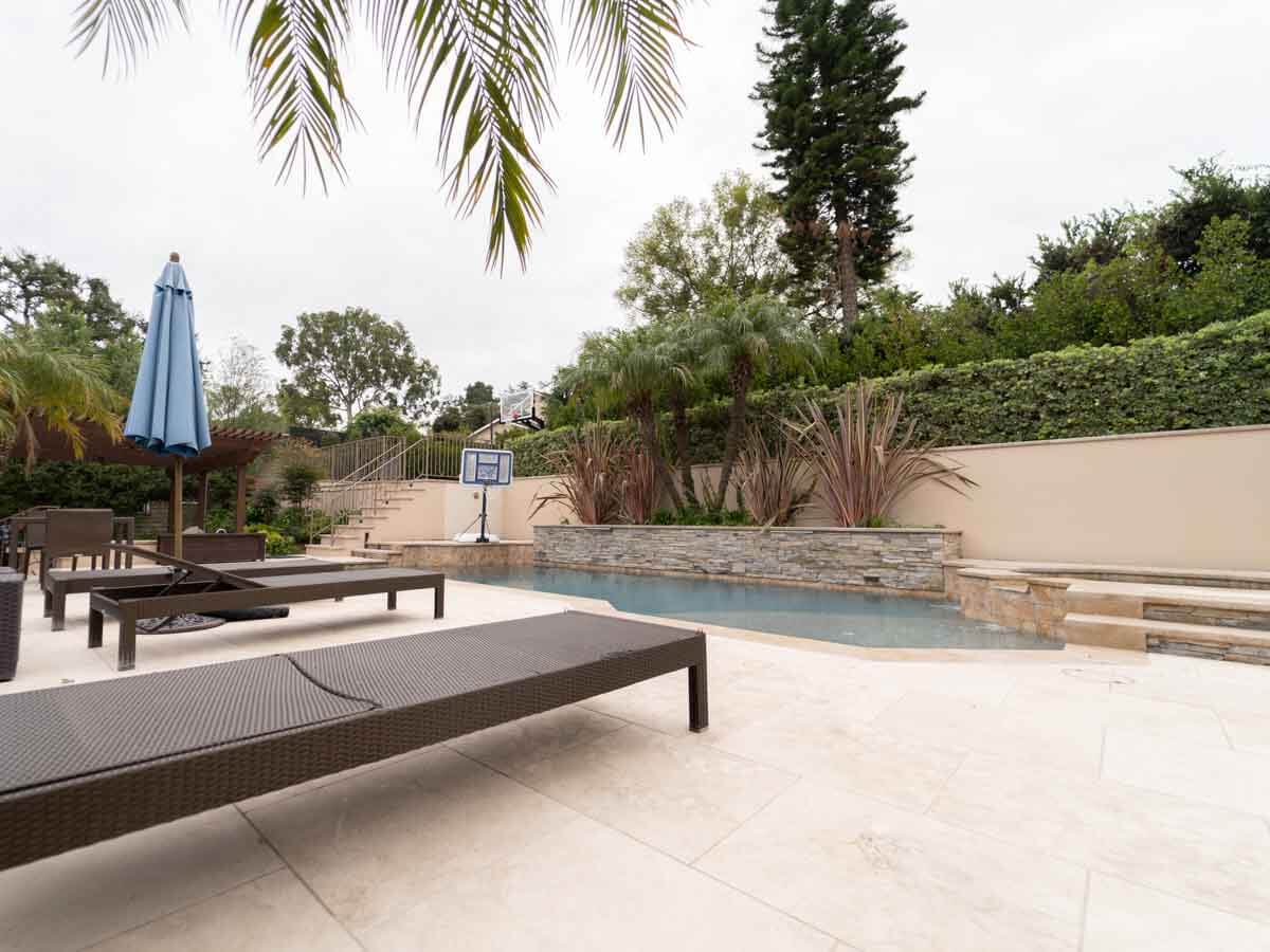 Wicker loungers under umbrellas on a travertine deck beside a raised stacked stone wall