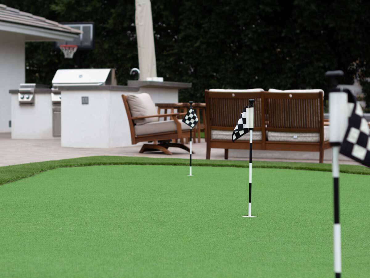 Putting green with checkered flags beside a white outdoor kitchen island and wood patio furniture