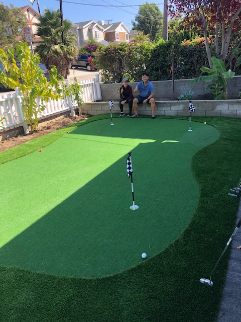Bright four-hole putting green beside a white picket fence with two people sitting on a low block wall