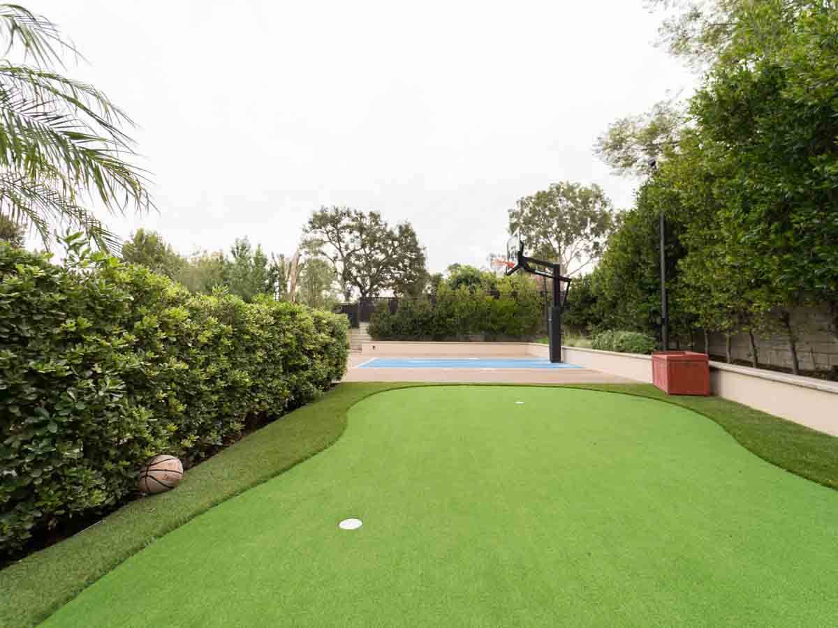 Backyard putting green alongside a tall hedge with a basketball court in the background